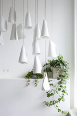 White porcelain chandelier shades hang at staggered heights against a white wall beside trailing green plants.