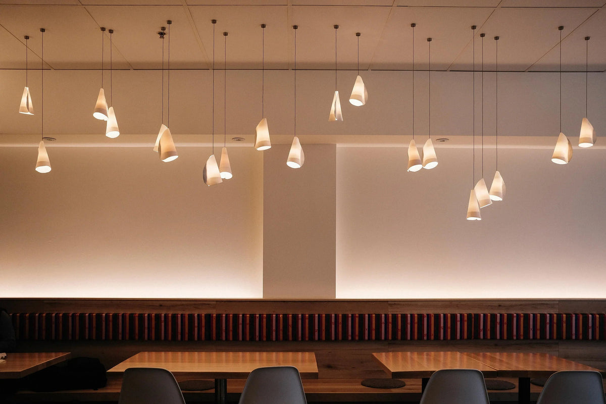 White porcelain chandelier shades hang at staggered heights above wooden dining tables and dark seating in a softly lit interior.