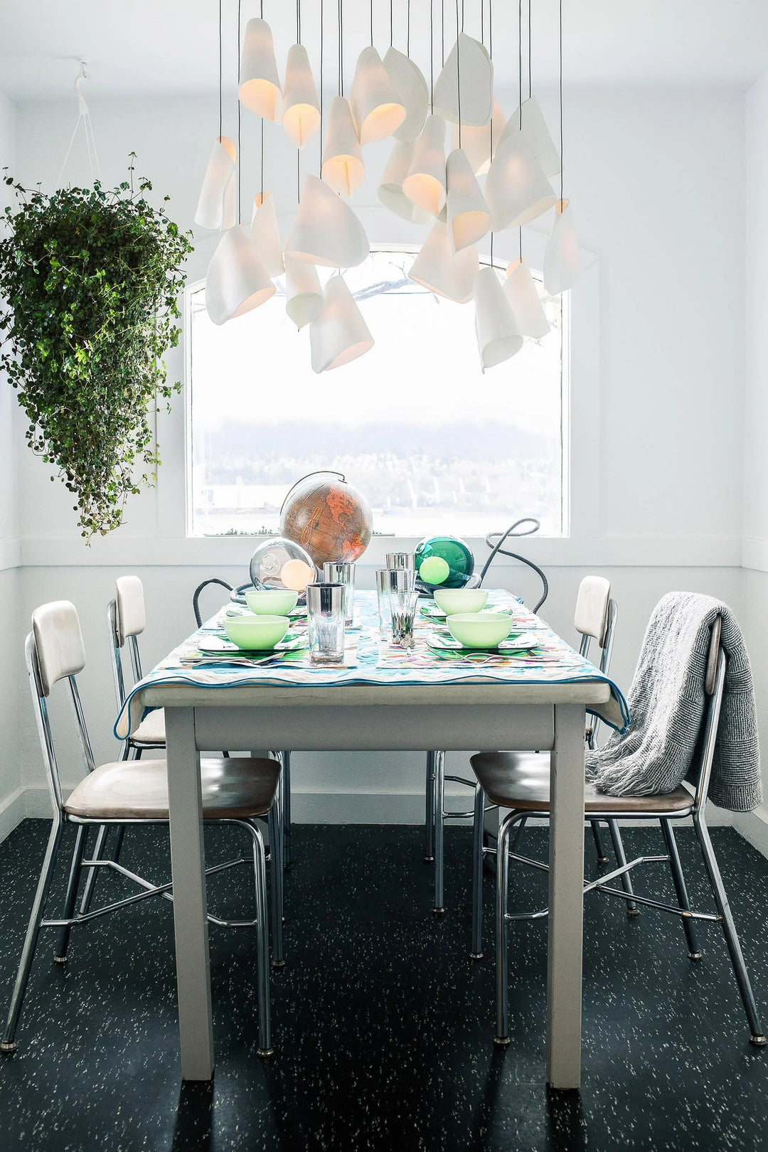 White porcelain chandelier with clustered folded shades hangs above a set dining table beside a window and trailing plant.