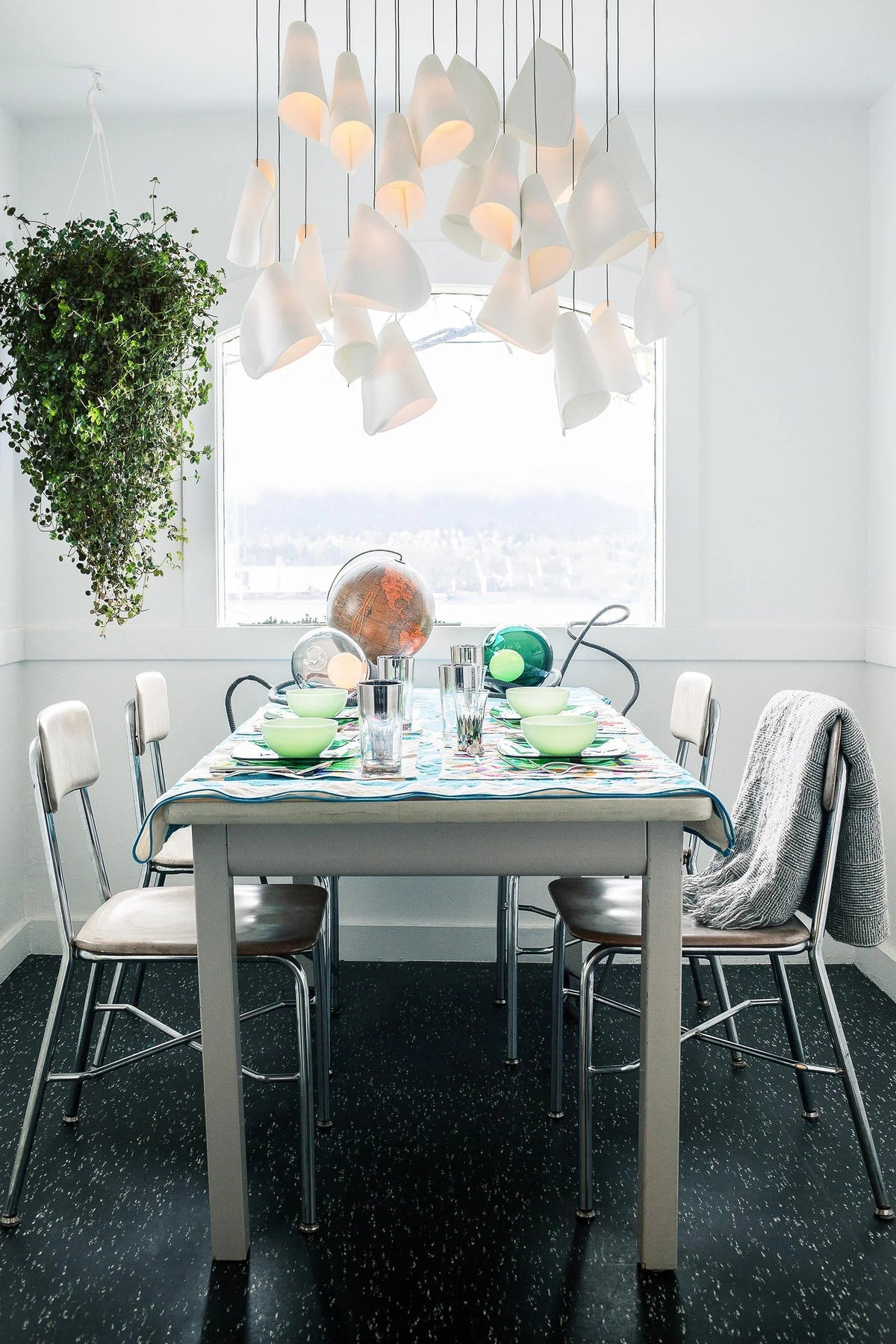 White porcelain chandelier with clustered folded shades hangs above a set dining table beside a window and trailing plant.