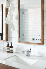 White porcelain chandelier with folded shades hanging beside a wood-framed mirror above a bathroom sink and white tiled wall.