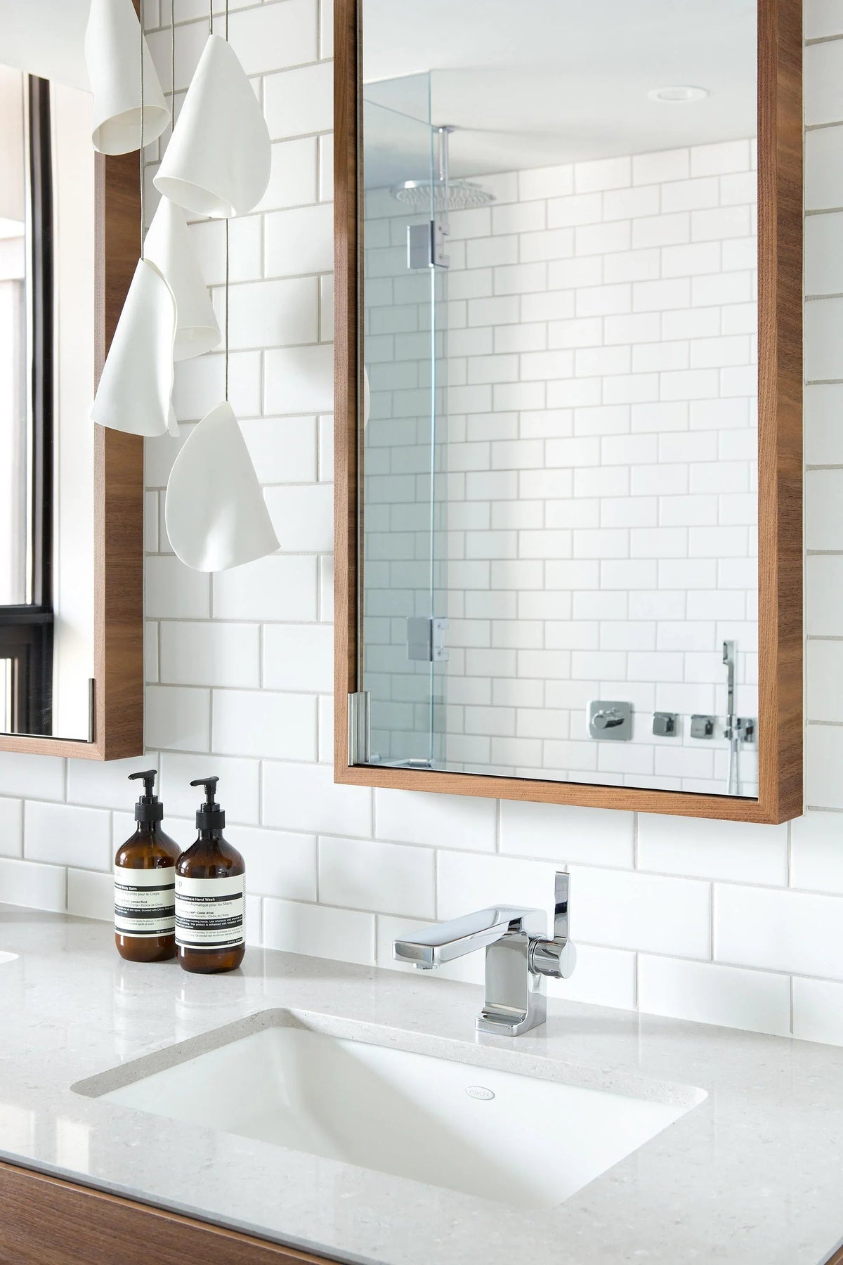 White porcelain chandelier with folded shades hanging beside a wood-framed mirror above a bathroom sink and white tiled wall.