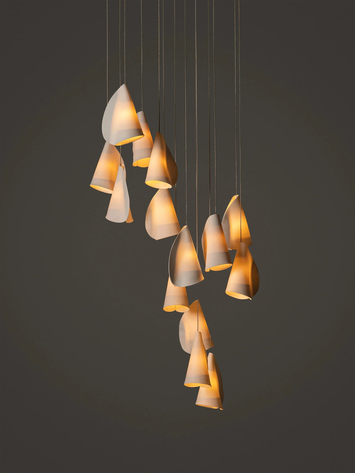 White porcelain chandelier with folded shades cascading at different heights and glowing warmly against a dark background.