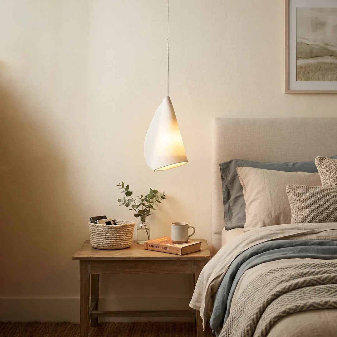 Chandelier shown with a single glowing white porcelain shade above a wooden bedside table beside an upholstered bed.