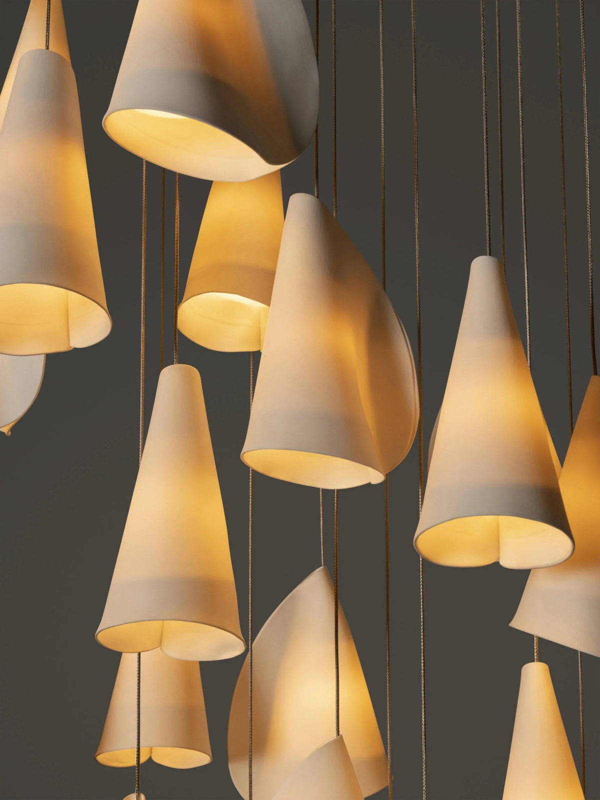 Close-up of white porcelain chandelier shades glowing golden against a dark gray background, with suspension cords visible.