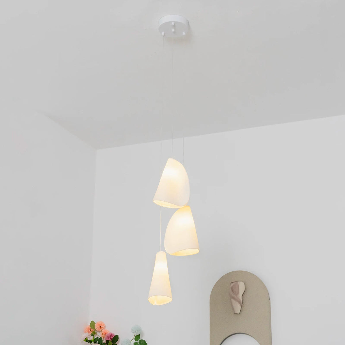 A three-light white porcelain chandelier hangs from a round ceiling canopy in a white room with flowers and wall decor.