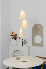 Three-light chandelier with glowing white folded shades hangs above a round white dining table near flowers and wall art.