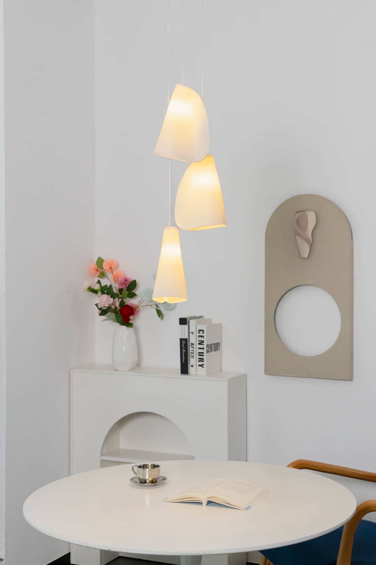 Three-light chandelier with glowing white folded shades hangs above a round white dining table near flowers and wall art.