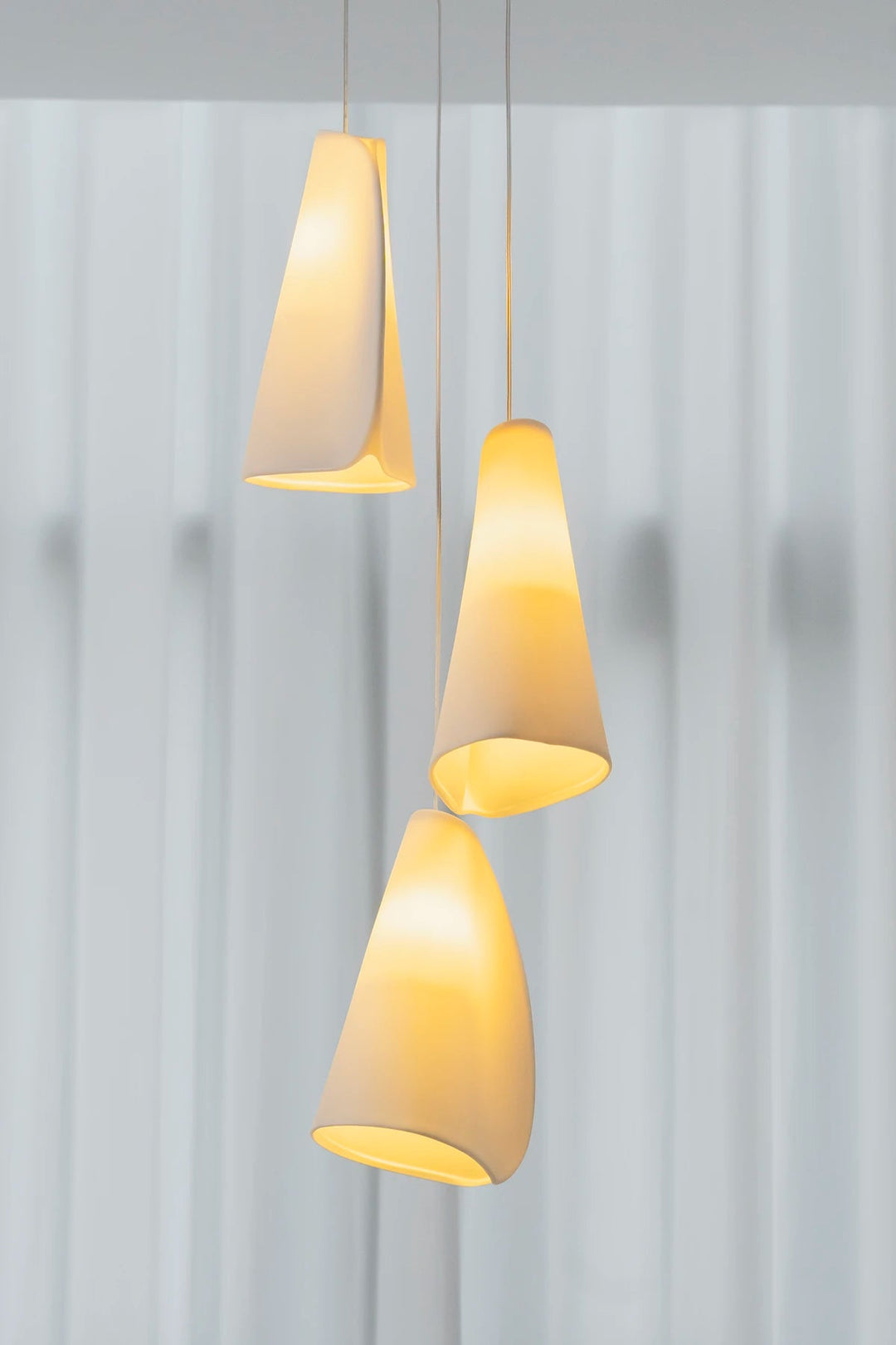 A three-light chandelier has folded white porcelain shades glowing yellow at staggered heights against pale curtains.
