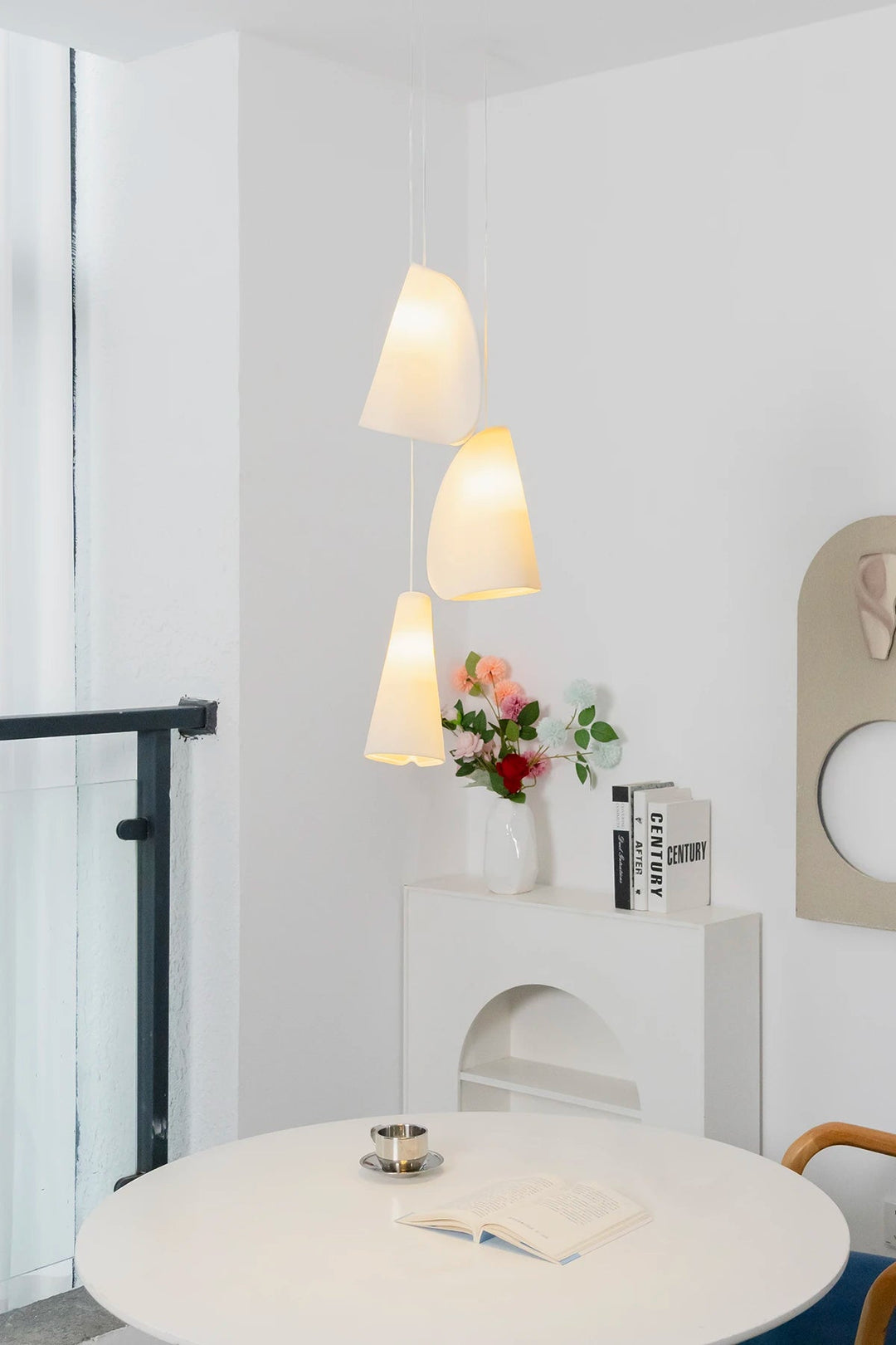 A modern three-light pendant chandelier with white fabric bell shades hangs over a round white table in a bright, minimalist room with white walls and a small white console shelf.