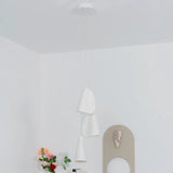 White porcelain chandelier with three folded shades hanging at staggered heights in a white room beside flowers and an arched mirror.