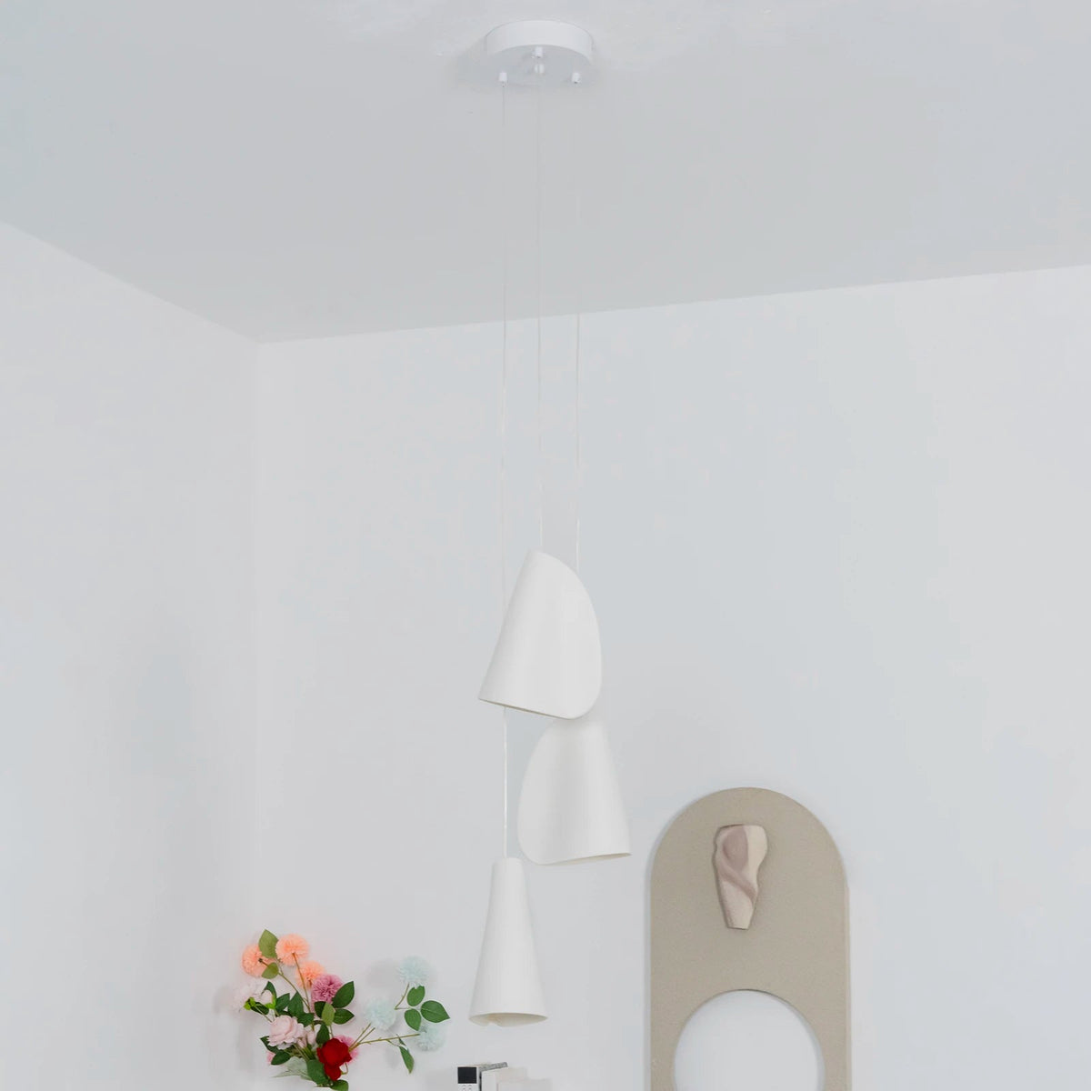 White porcelain chandelier with three folded shades hanging at staggered heights in a white room beside flowers and an arched mirror.