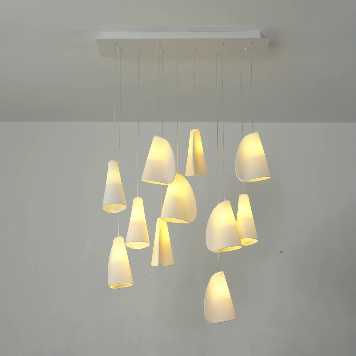 A modern multi-light pendant chandelier with multiple tapered white shades of varying sizes, suspended from a rectangular ceiling plate against a plain light gray wall.