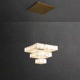 A modern multi-tiered geometric chandelier with white marble-look rectangular shades suspended from a square gold-toned ceiling canopy against a dark gray wall.