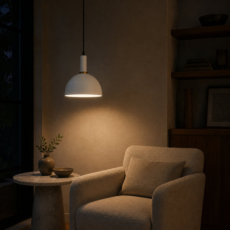 A modern pendant light with a white dome shade and gold-toned trim hangs over a beige armchair in a dimly lit, warm-toned living room corner.
