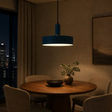 A modern cylindrical pendant light with a dark teal-blue shade and a warm white glowing bulb, hanging over a round wooden dining table in a warmly lit, contemporary room.