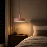 A modern pink pendant light with a cylindrical shade and black cord hangs over a bed in a softly lit bedroom. The lamp glows warm yellow light, casting a glow on a textured beige wall and a wooden nightstand with a small vase and plant.