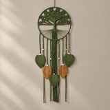 A green macrame tree of life wall hanging with five hanging leaf charms, including three tan leaves and two green leaves, displayed against a neutral beige wall with soft diagonal shadows.