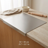 A rectangular stainless steel cutting board with rounded corners and raised edges, shown resting on a light wood countertop. A beige cloth, a small bowl, and a wooden rolling pin are nearby, and text at the bottom reads 50 x 40 cm / 19.