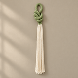 A decorative hanging tassel with a knotted sage green top and a long cream tassel, shown against a warm beige wall with soft window-shadow lighting.