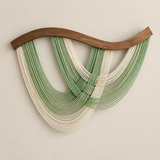 A modern wall art sculpture featuring a wavy wooden top bar and multiple layered strands in green and cream hanging in a fan-like arrangement against a plain beige wall.