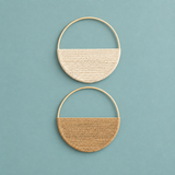 Two semi-circular hoop earrings with woven textured halves, one in cream and one in tan, displayed on a solid teal background.