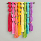 A colorful wall hanging made of multiple vertical strands of yarn tied in round beads, hanging from a dark horizontal rod against a plain light gray wall.