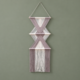 A geometric macrame wall hanging with a dusty lavender and white color-blocked pattern, suspended from a wooden dowel against a muted sage green wall.