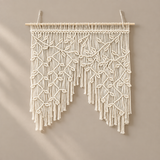 Cream macrame wall hanging with intricate woven patterns and long fringe, suspended from a light wooden dowel against a warm beige wall.