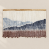 A large woven wall hanging with vertical strands that form a mountain-like abstract pattern in cream, blue, and taupe-brown, suspended from a light wood rod against a warm beige wall.