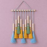 A bohemian-style wall hanging decoration featuring a light wood dowel, light blue hanging cords, white wooden beads, and tassels in light blue, sage green, and mustard yellow, set against a solid dusty pink background.