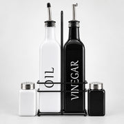 A set of kitchen bottles and shakers on a black metal stand against a light gray background. The set includes a tall white bottle labeled 'OIL', a tall black bottle labeled 'VINEGAR', a small white shaker, and a small black shaker with silver metal tops.