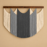 A bohemian-style macrame wall hanging with a dark brown wooden top bar, featuring vertical fringe strands in dark gray, medium gray, and cream, set against a solid beige wall.