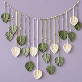 A decorative hanging garland made of woven palm-leaf shapes in sage green and cream, suspended from a string with small round beads, against a solid lavender background.