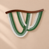 A modern wall hanging sculpture featuring three interlocking U-shaped strands in shades of forest green, sage green, and off-white, suspended from a curved dark brown wooden bar against a warm beige background.