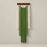 A vertical woven wall hanging with a dark wood top bar, featuring a central green fringe panel flanked by two cream fringe panels, displayed against a plain light gray wall.