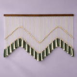 A bohemian-style wall hanging with a wooden top bar, white vertical cords, gold bead accents, and green and cream tassels arranged in a zigzag pattern against a solid lavender wall.