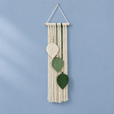 A bohemian-style macrame wall hanging with cream cotton cord, wooden dowel, and three hanging leaf shapes in cream and dark green, shown against a dusty blue wall.