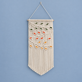 A cream macrame wall hanging with a woven tree design featuring orange, yellow, and green leaves, suspended from a light wood dowel against a solid blue background.