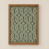 A framed macrame wall hanging with an intricate woven geometric pattern in sage green rope, enclosed in a light brown wooden frame and mounted on a warm beige wall.