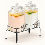 Two large glass beverage dispensers with lids and metal spouts, displayed on a black metal stand. The left dispenser contains a pale pink drink with lemon slices, and the right dispenser contains a clear drink with citrus slices and mint leaves.