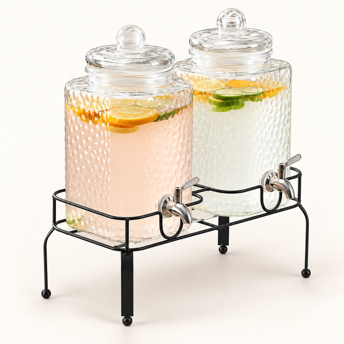 Two large glass beverage dispensers with lids and metal spouts, displayed on a black metal stand. The left dispenser contains a pale pink drink with lemon slices, and the right dispenser contains a clear drink with citrus slices and mint leaves.