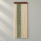 A vertical woven wall hanging with a natural wood top bar, featuring vertical cream and sage green cords with a central leafy vine motif, displayed against a light gray wall.