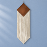 A geometric boho wall hanging featuring a warm brown wood diamond top and long cream cotton fringe, suspended against a light blue wall.