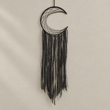 A black crescent moon dreamcatcher with intricate woven webbing and long dangling fringe, hanging against a plain beige wall.