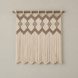 A bohemian-style macrame wall hanging with intricate geometric diamond patterns and long fringe, suspended from a natural wood dowel against a solid light gray wall.