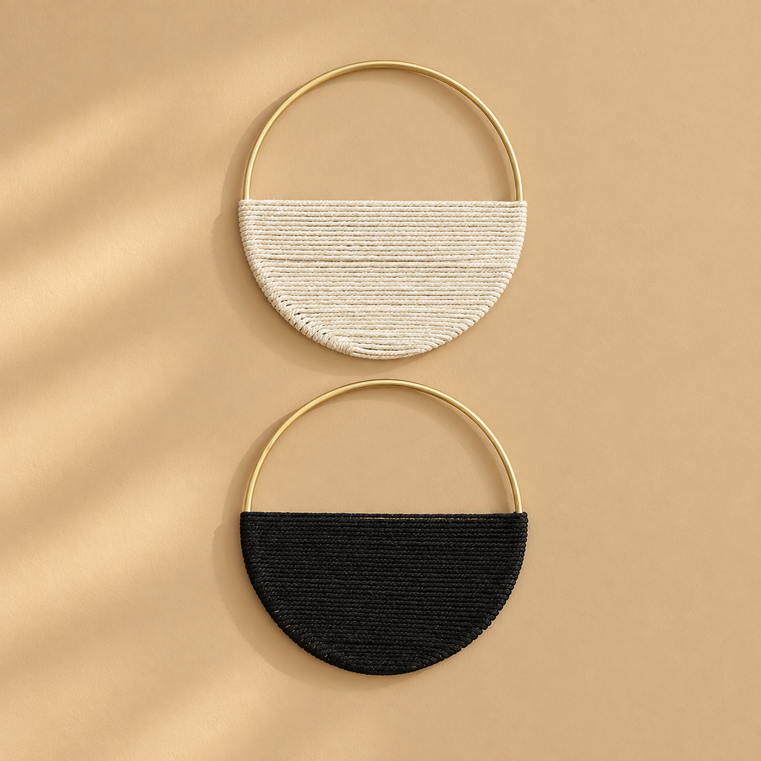 Two semi-circular woven half-moon handbags with gold-tone metal hoops, one in off-white and one in black, displayed on a warm beige background with soft diagonal shadows.