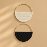 Two semi-circular woven half-moon handbags with gold-tone metal hoops, one in off-white and one in black, displayed on a warm beige background with soft diagonal shadows.