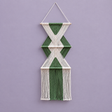 A geometric macrame wall hanging with alternating green and off-white woven cords, suspended from a wooden dowel against a solid lavender-purple background.