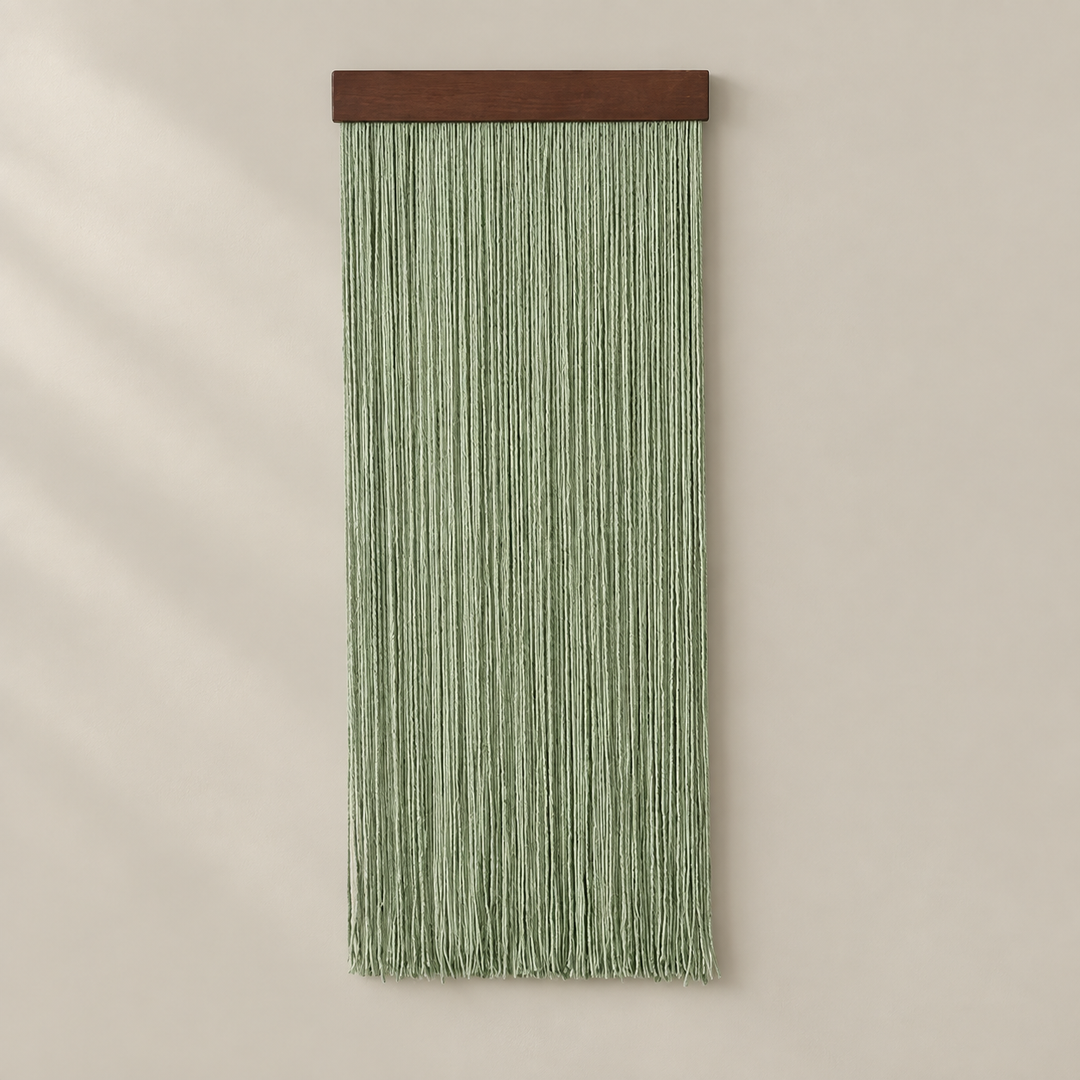 A vertical sage green macrame wall hanging with a dark brown wooden top bar, displayed against a warm beige wall with soft diagonal shadows.